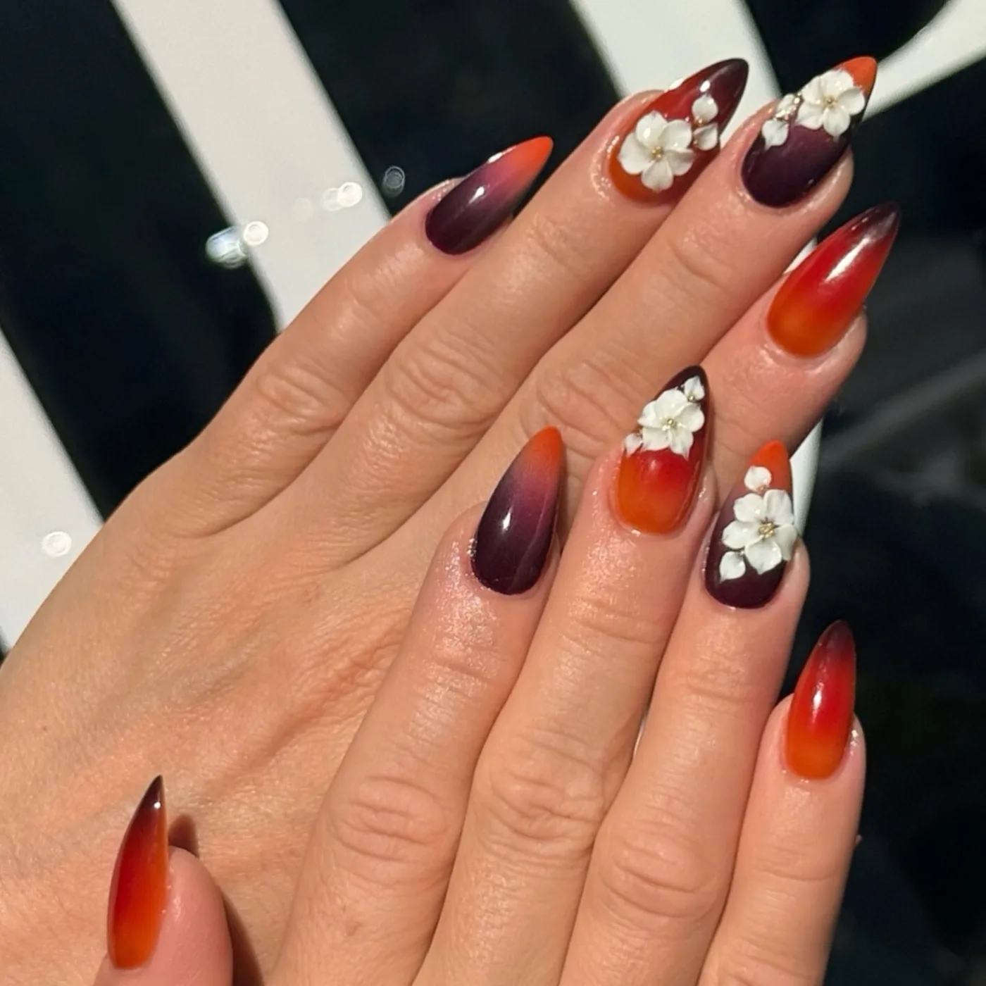 Mandelförmige Nägel in Bordeaux und Orange mit weissen Blüten, im Hintergrund das Della Logo am Schaufenster